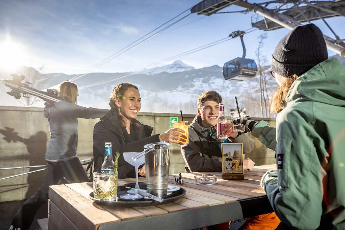 Four people raising their drinks in the outdoor area of the e-tron energy bar as an Eiger Express gondola passes by.